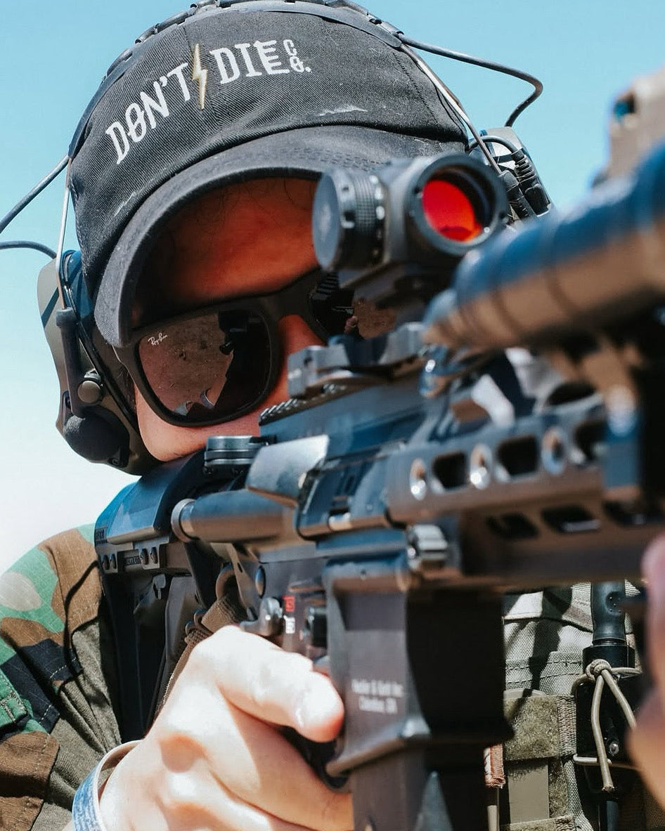 Person in tactical gear with a rifle, wearing a 'Don't Die' cap.